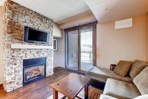A living room with a stone fireplace, a flat-screen TV mounted above it, a sofa, and a window with blinds. - Beautiful studio w/private balcony & shared pool, hot tubs, fitness & game room (Steamboat Springs)