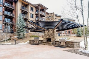 A covered outdoor grill area with stone pillars, surrounded by a courtyard with a building in the background. - Luxurious condo w/ private balcony & shared pool, hot tubs, game room & fitness (Steamboat Springs)