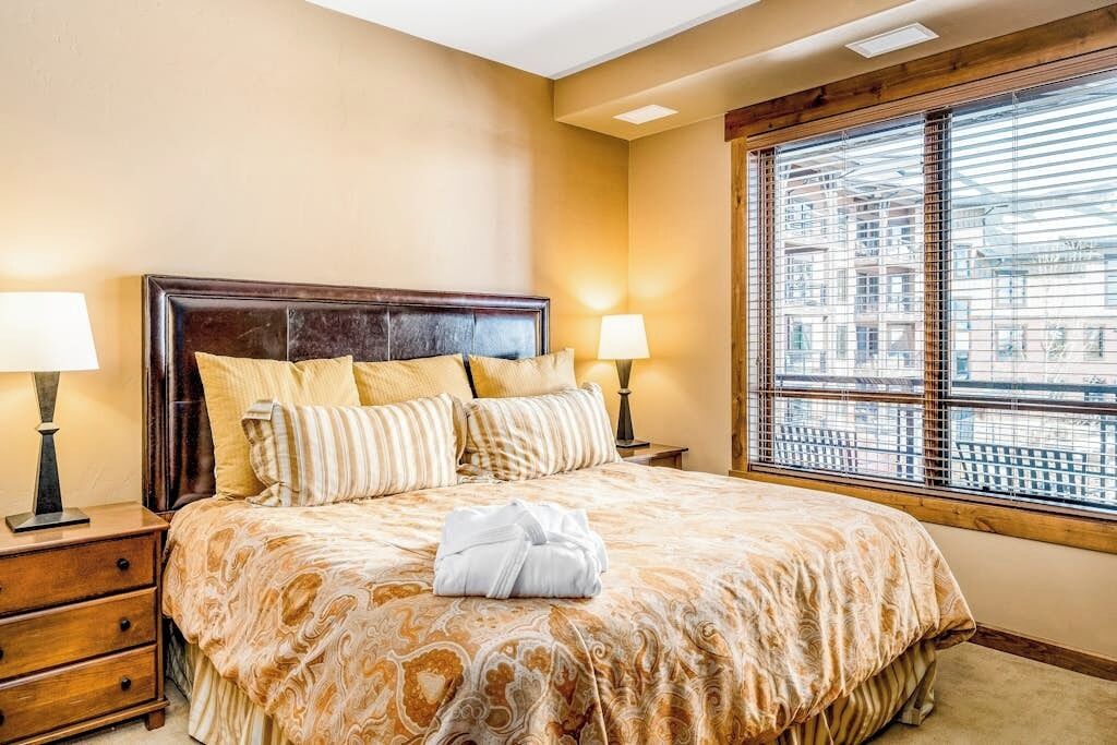 A hotel room with a large bed, two bedside tables with lamps, a window with blinds, and a view of buildings outside.