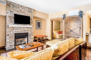 A living room with a stone fireplace, a sofa, a coffee table, and a sliding barn door. - Luxurious condo w/ private balcony & shared pool, hot tubs, game room & fitness (Steamboat Springs)