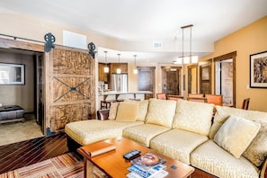 A living room with a sectional sofa, a coffee table, and a wooden wall with a hanging rack. - Luxurious condo w/ private balcony & shared pool, hot tubs, game room & fitness (Steamboat Springs)