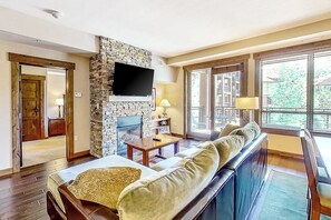 Ein Wohnzimmer mit Kamin aus Stein, einem Flachbildfernseher, einer Couch und großen Fenstern.