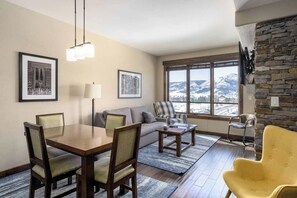 Ein Wohnzimmer mit Esstisch, Sofa und Stühlen, einem großen Fenster mit Blick auf die Berge und einer Wand aus Naturstein.