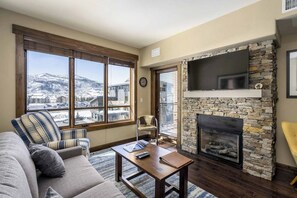 Ein Wohnzimmer mit Kamin aus Stein, einem darüber montierten Fernseher, einer Couch und Blick auf eine verschneite Landschaft durch große Fenster.