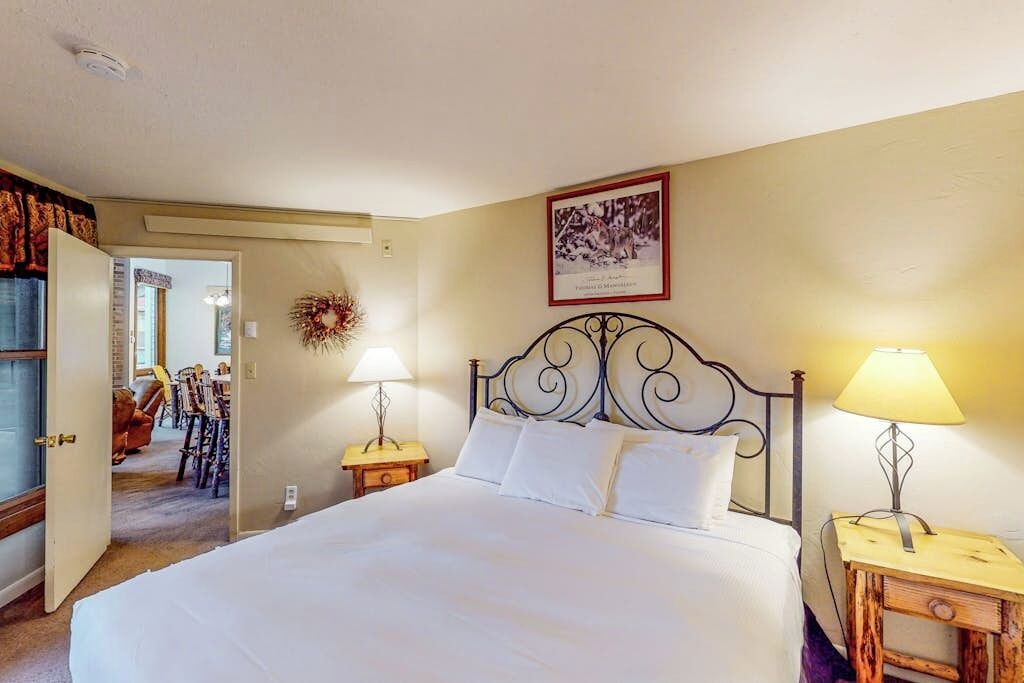 Quarto com cama, duas luminárias de cabeceira, um quadro na parede e uma porta que dá para outro cômodo.