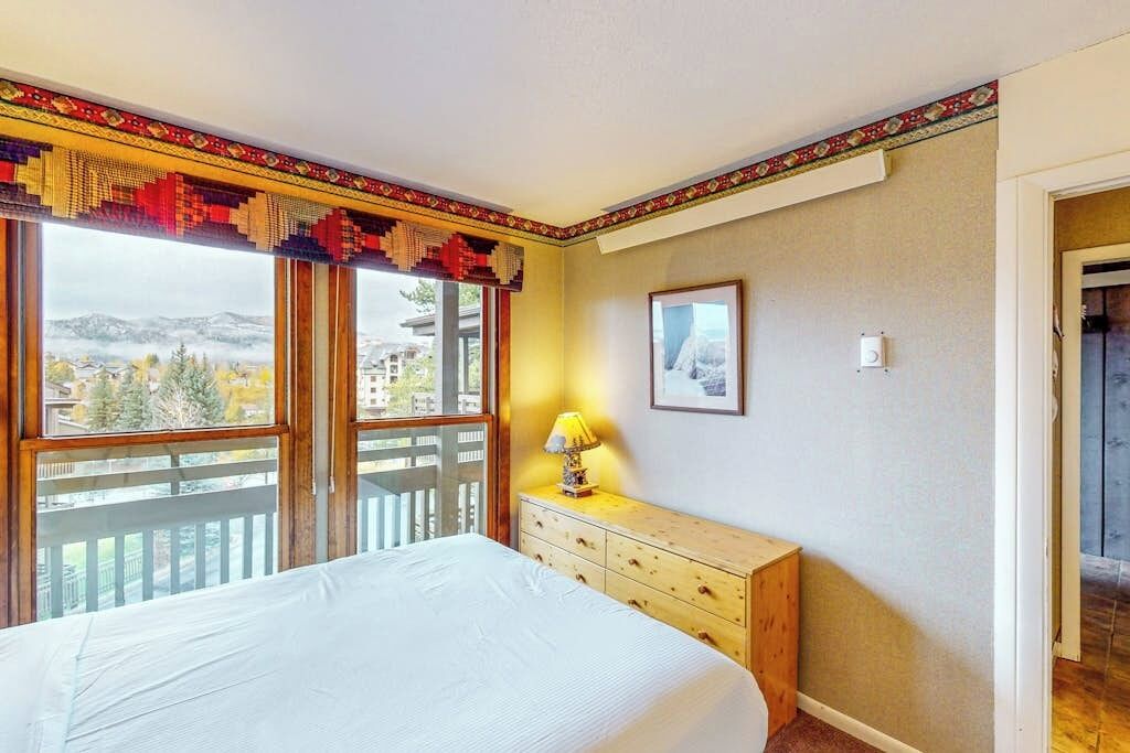 Une chambre à coucher comprenant un lit, une commode, une lampe, un tableau encadré et une fenêtre donnant sur un balcon et des immeubles.