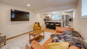 A living room with a sofa, coffee table, and TV. There is a kitchen in the background with a dining area. - King Bed, 75 min Banff, AC Backyard Chickens (Calgary)