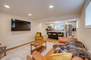 Ein Wohnzimmer mit Sofa, Couchtisch und Fernseher. Im Hintergrund ist eine Küche mit Essbereich zu sehen.