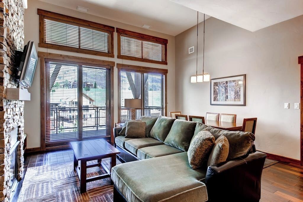 A living room with a sectional sofa, a TV mounted on a stone wall, and large windows with blinds.