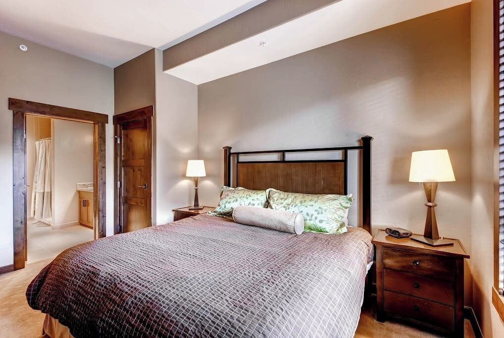 Une chambre à coucher avec un grand lit, deux tables de chevet, une tête de lit en bois et une porte donnant sur une autre pièce.