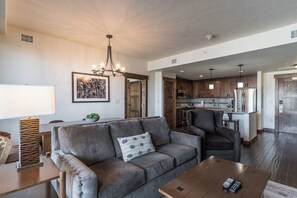 Ein modernes Wohnzimmer mit einer Couch, einem Couchtisch und einer Chandelieber. Im Hintergrund ist eine Küche mit Holzschränken und Edelstahlgeräten zu sehen.