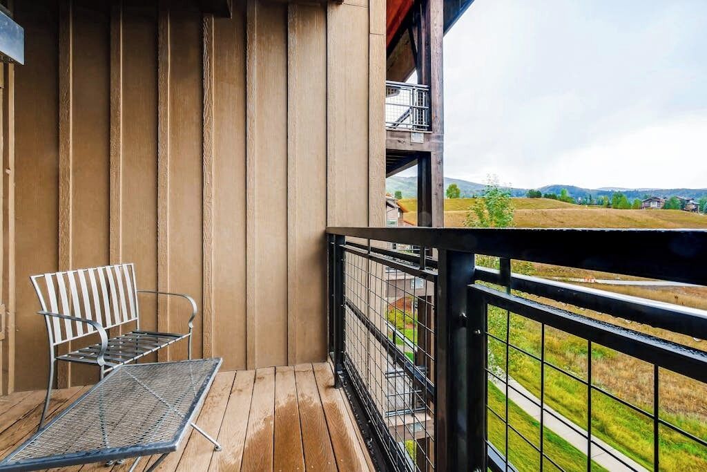 Un balcone con una sedia e un tavolo in metallo, pannelli in legno, una ringhiera metallica e vista su un paesaggio verde.