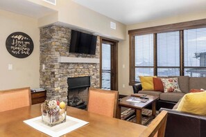 Ein Wohnzimmer mit Kamin aus Stein, einem darüber montierten Fernseher, einer Couch mit bunten Kissen und einem Essbereich mit Tisch und Stühlen.