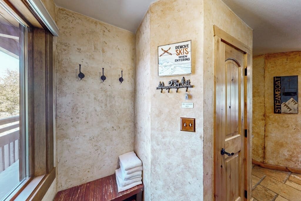 A rustic bathroom with a wooden bench, a towel rack, a sign instructing to remove skis, and a door labeled 'STEAMBOAT'.