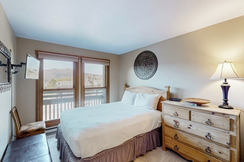 Une chambre à coucher comprenant un lit, une commode, une lampe, un élément de décoration murale et une fenêtre donnant sur un paysage agréable.