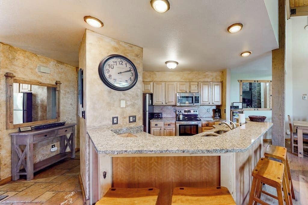 Une cuisine dotée de plans de travail en granit, de meubles en bois, d’un îlot central et d’une horloge murale.