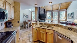 Une cuisine dotée de plans de travail en granit, de meubles en bois, d’un lave-vaisselle, d’une cuisinière et d’une grande fenêtre donnant sur une forêt.