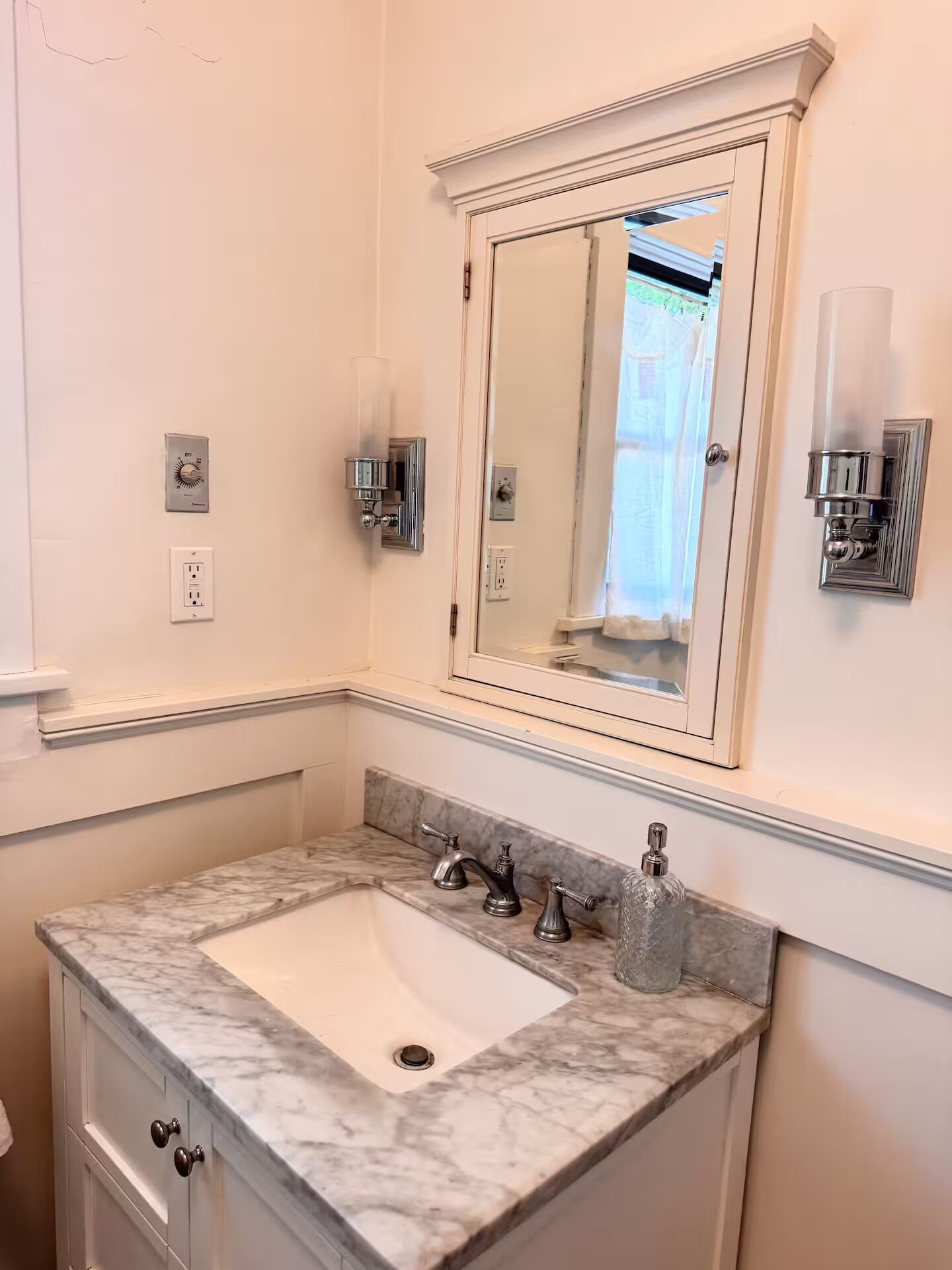 A bathroom with a marble sink, a large mirror, a wall-mounted light, and a thermostat.