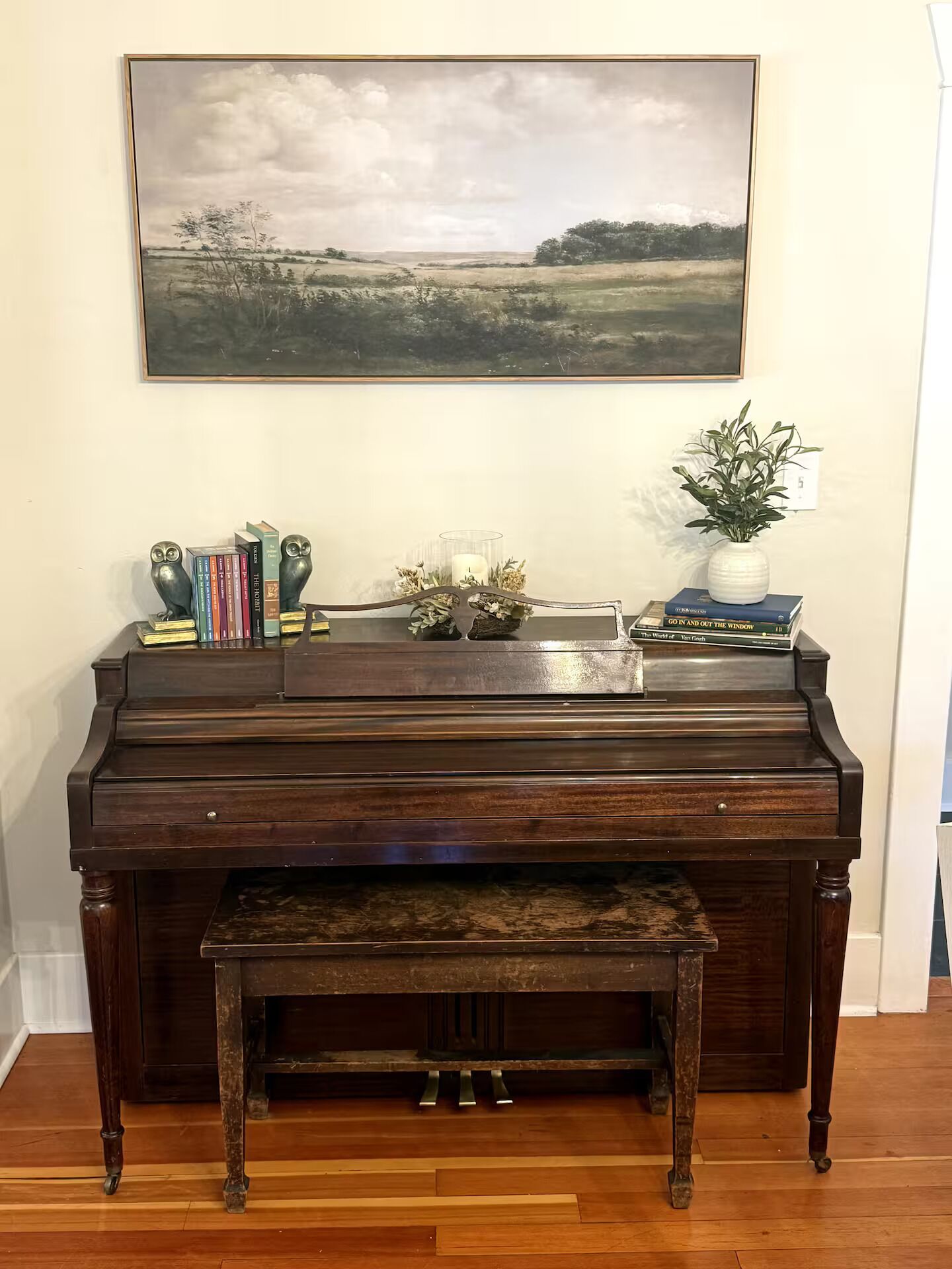 Een vleugelpiano met een bankje, een lijstwerk landschap en een plant in een pot op een tafel.