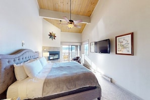 A bedroom with a large bed, a ceiling fan, a mounted TV, and a window with a view. - Large updated condo with full kitchen & private balcony - near resort trails (Steamboat Springs)