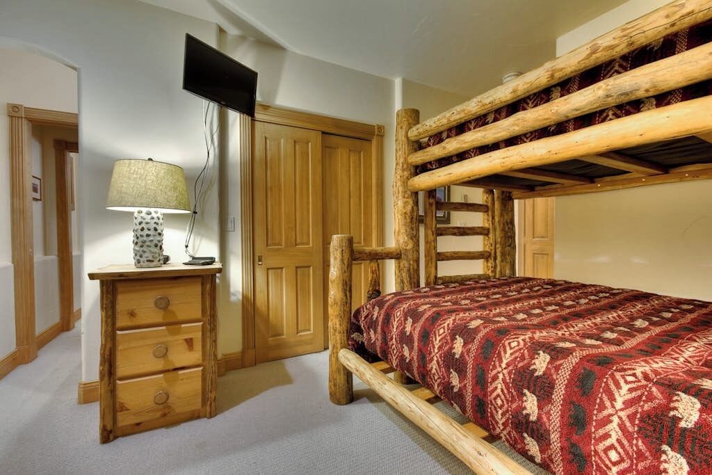 Ein Etagenbett mit gemustertem Bettbezug, ein Holztisch mit Schubladen, eine Lampe und ein Fernseher an der Wand.