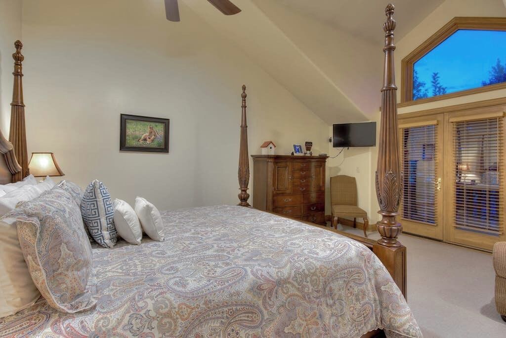 Een slaapkamer met een hemelbed, een bureau, een stoel en een groot raam met jaloezieën.
