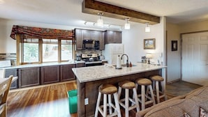 A kitchen with dark cabinets, a central island with stools, a large window, and a ceiling with exposed beams. - Intimate condo with fireplace, washer/dryer, outdoor pool & hot tub (Steamboat Springs)