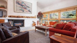 A living room with a fireplace, a sofa, a coffee table, and a large window with a view of greenery. - Kutuk 303 - Spacious, top-floor condo near ski area w/ mountain views plus shared hot tubs (Steamboat Springs)