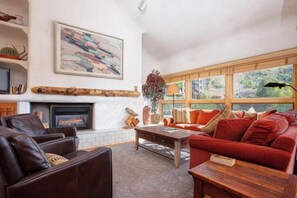 A living room with a fireplace, a sofa, a coffee table, and a large window with a view of greenery. - Spacious, top-floor condo near ski area w/ mountain views plus shared hot tubs (Steamboat Springs)