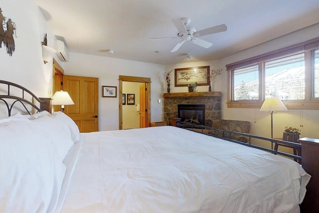 A bedroom with a bed, a fireplace, a ceiling fan, and a window.