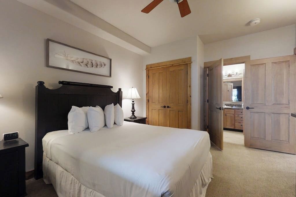 Quarto com cama, mesa de cabeceira, abajur, ventoinha no teto e porta de madeira.