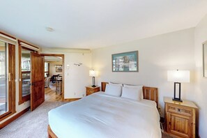 Quarto com cama grande, mesas de cabeceira de madeira, um quadro na parede e uma porta que dá para outro cômodo.