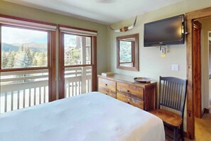 Ein Schlafzimmer mit Bett, Schminktisch, Stuhl, Fernseher und einem Balkon mit Blick auf Bäume und Gebäude.