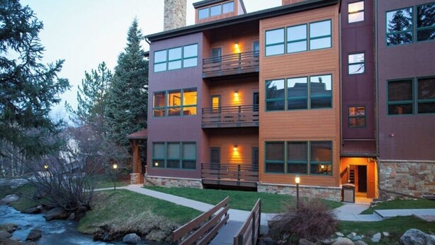A multi-story building with balconies, a stone chimney, and a wooden deck over a stream. - Kutuk 212 - Recently remodeled condo close to the base area w/ shared hot tubs & firepit (Steamboat Springs)