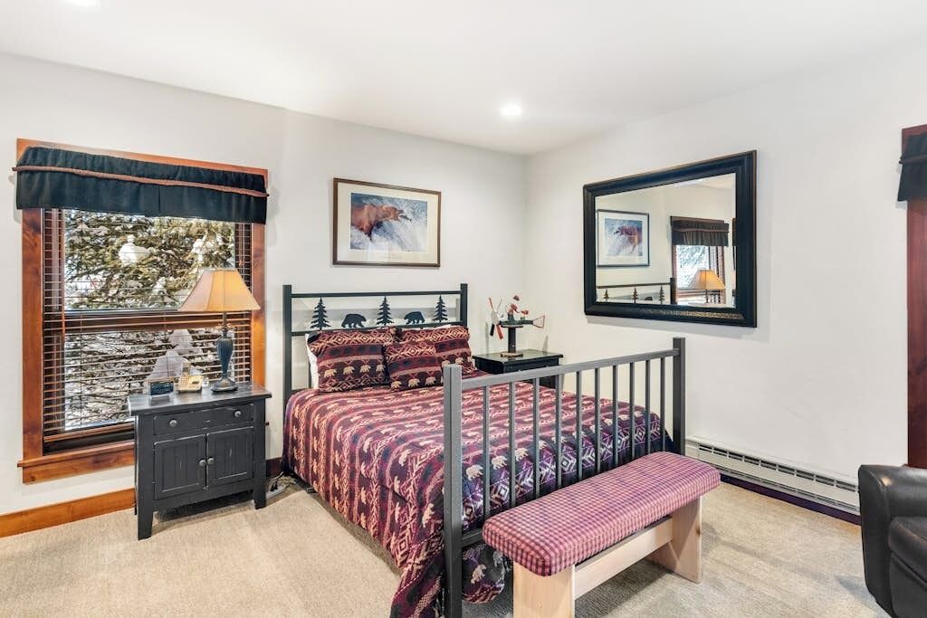 A bedroom with a bed, a bedside table, a bench, a window with blinds, a mirror, and a framed picture on the wall.