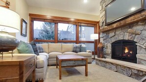 A cozy living room with a stone fireplace, a sofa with cushions, a wooden coffee table, and a TV mounted above the fireplace. - Kutuk 212 - Recently remodeled condo close to the base area w/ shared hot tubs & firepit (Steamboat Springs)