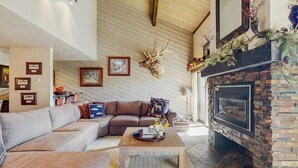 A cozy living room with a stone fireplace, a large deer head mounted on the wall, and a sectional sofa. - Top-level condo close to summer fun with grills, hot tubs & parking (Steamboat Springs)