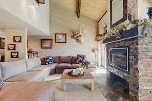 A cozy living room with a stone fireplace, a large deer head mounted on the wall, and a sectional sofa. - Top-level condo close to summer fun with grills, hot tubs & parking (Steamboat Springs)