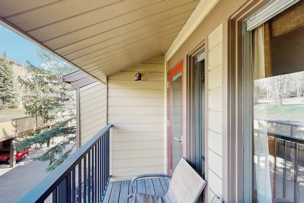 A covered balcony with a chair, a door with a red frame, and a view of a parking area and trees.