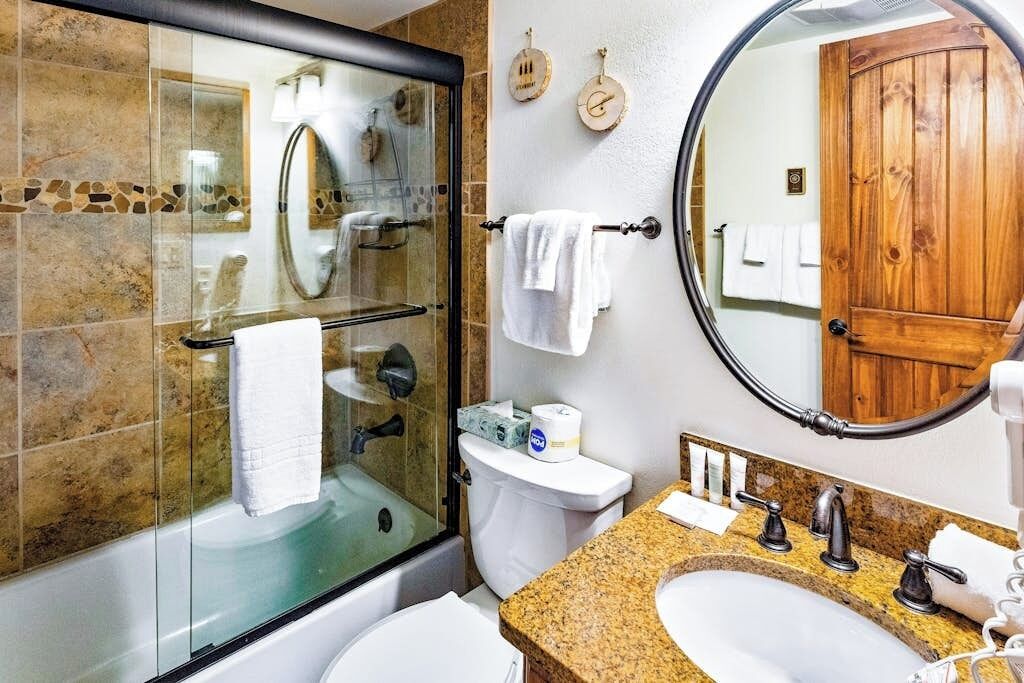 Un baño con ducha envuelta en vidrio, inodoro blanco, lavabo con encimera de granito y puerta de madera.