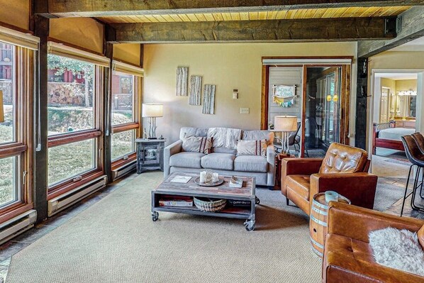 A spacious living room with a sofa, coffee table, and leather armchairs. Large windows offer views of the outdoors.