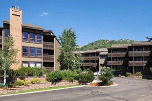 A multi-story hotel with balconies, a parking lot, and a hilly landscape in the background.