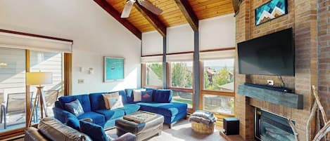 Una sala de estar amplia con un sofá seccional azul, una chimenea, un televisor de pantalla plana y un ventilador de techo.