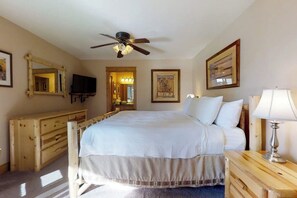 Ein Schlafzimmer mit einem Bett, einem Deckenventilator, einem Fernseher, einer Holzkommode, einer Lampe und gerahmten Kunstwerken an den Wänden.