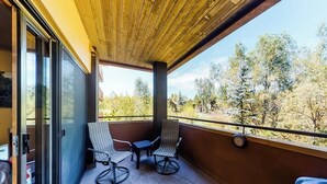 A balcony with two chairs and a table, a clock on the wall, and a view of trees. - Canyon Creek A105 - Family-friendly condo w/mountain views, balcony, shared hot tub & outdoor pool (Steamboat Springs)