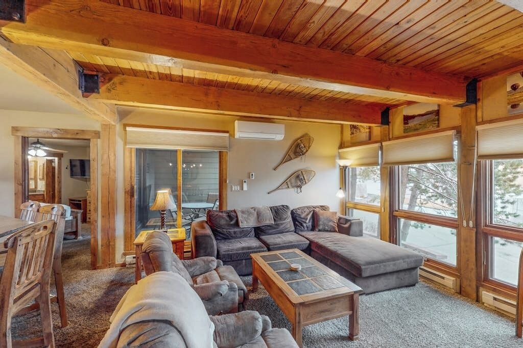 Un salon chaleureux avec un plafond en bois, un canapé d’angle, une table basse et de grandes fenêtres.