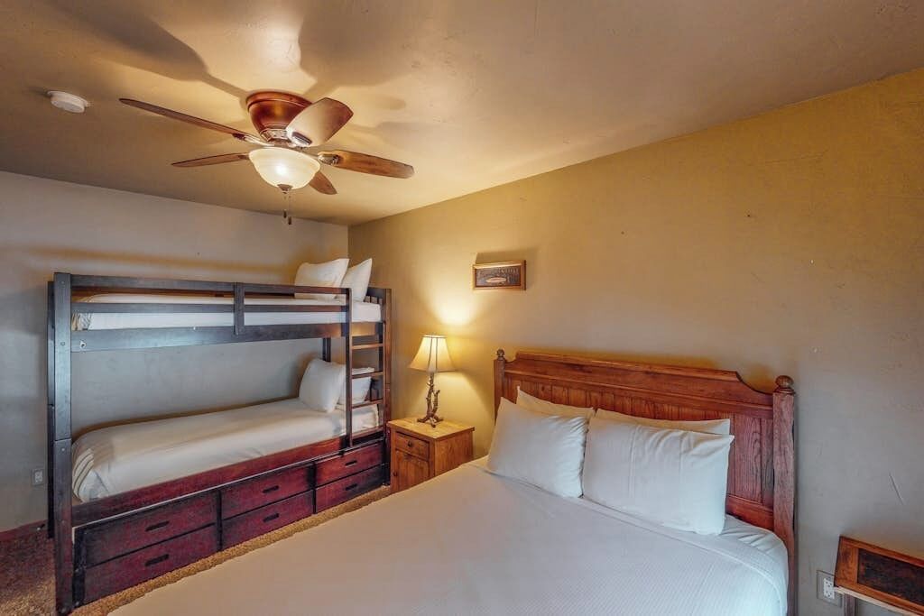 Een kamer met een stapelbed, een enkel bed, een houten hoofdbord, een nachtkastje met een lamp en een plafondventilator.