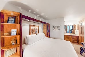 Ein Schlafzimmer mit einem großen Bett, Holzregalen mit gerahmten Bildern, einer kleinen Kommode und Blick ins angrenzende Badezimmer.