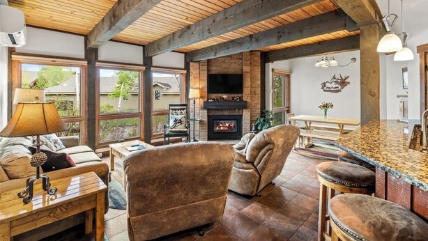 A cozy living room with a fireplace, wooden ceiling beams, a sofa, and a bar area. - Steamboat home with washer/dryer, pools & hot tubs - near ski lifts (Steamboat Springs)