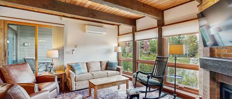 A cozy living room with a fireplace, leather furniture, and large windows offering a view of the outdoors.